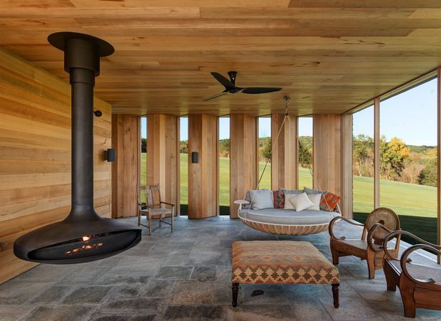A warm, open-sided sunroom or living space with full-height glazing overlooking a landscaped rural setting. The room features a dramatic suspended black wood-burning fireplace, timber-clad walls and ceiling, large-format grey stone flooring, a suspended woven daybed, wicker lounge chairs, and an upholstered patterned ottoman. The design blends natural materials with mid-century-inspired furnishings and strong indoor-outdoor connection.