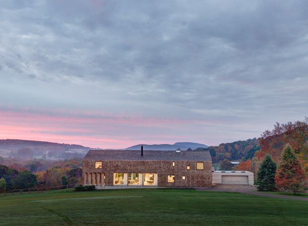 Exterior architectural photograph of a low, elongated rural residence at dusk. The house has a steep pitched roof, dark shingle or weathered-cladding surfaces, warm illuminated windows, a glazed central living area, and an attached garage. It sits in a broad manicured lawn with wooded hills and autumn vegetation in the background beneath a dramatic cloudy sky.