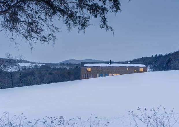 A low, modern rectangular house set in a snow-covered rural or mountainous landscape at dusk. The dark grey facade has a shallow pitched or nearly flat roof blanketed with snow, a chimney, and warm illuminated windows. Bare woodland, rolling hills, and overhanging branches frame the isolated dwelling.