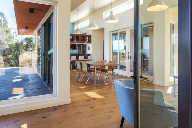 Bright contemporary open-plan dining and living area viewed through glazed doors, with warm timber flooring, a wooden dining table and chairs, sculptural pendant lights, built-in shelving, and extensive floor-to-ceiling glazing opening toward an outdoor terrace. A blue upholstered armchair is visible in the foreground, and the space has a strong indoor-outdoor connection.