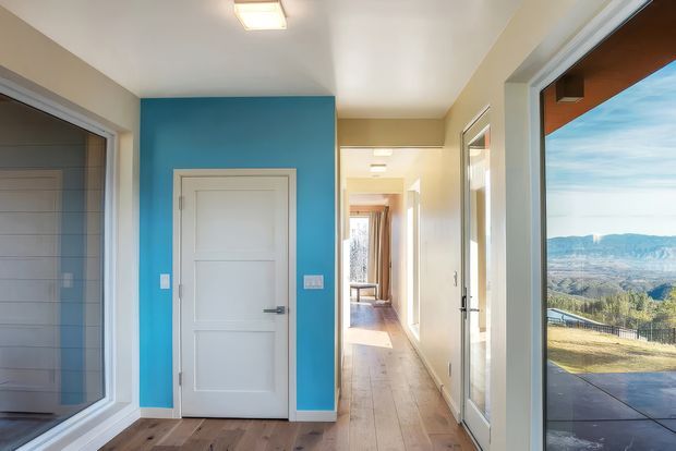 Bright contemporary hallway with warm timber flooring, white walls and doors, and a vivid blue feature wall around a paneled interior door. Glazed openings on both sides provide views into adjacent rooms and out toward a scenic mountainous landscape and terrace. Recessed ceiling lighting and clean-lined trim create a modern, airy circulation space.