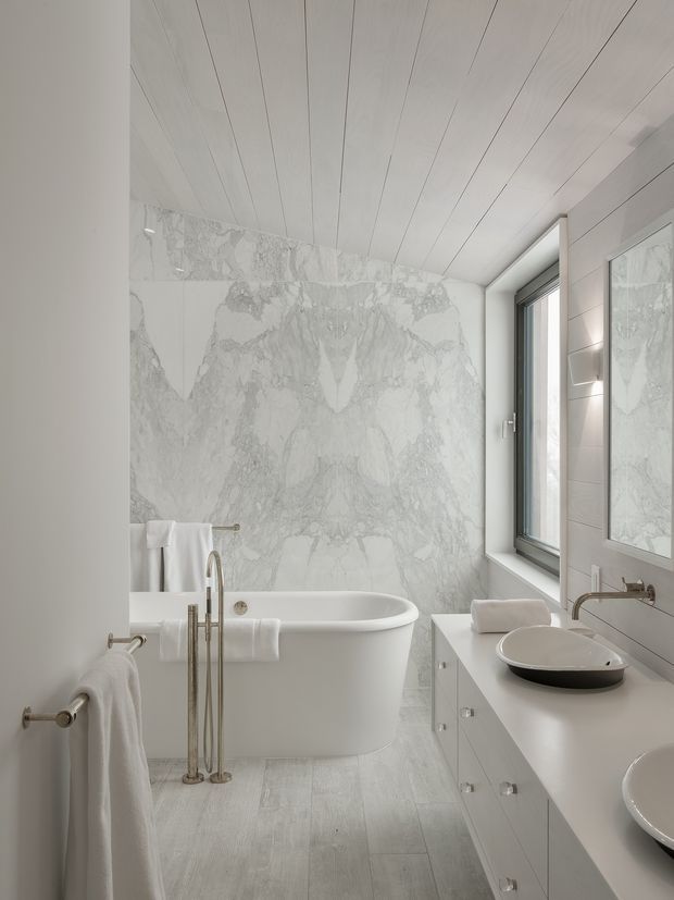 Bright, refined contemporary bathroom featuring a freestanding white bathtub, wall-mounted vanity with vessel basins, brushed metal tapware, a large marble-look feature wall, pale tiled flooring, white wall panelling, and a tall window. The room has a clean, light monochromatic palette with subtle grey veining.