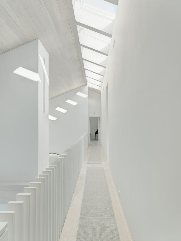 A bright, minimalist interior hallway or landing overlooking a stairwell. White walls, pale timber ceiling boards, white stair balustrades, and a long skylight strip create a highly luminous, narrow circulation space. A pale runner extends along the floor, with strong geometric daylight patterns cast through the roof glazing.