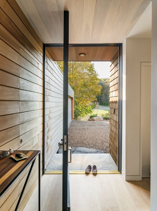 A refined residential entryway viewed from inside, with a full-height black-framed glazed door opening to a gravel path and landscaped garden. The entry is enclosed by pale timber wall and ceiling panelling, with matching light timber flooring, a slim black-framed console, recessed ceiling lighting, and a built-in white cupboard. A narrow covered threshold and exterior timber-clad walls are visible beyond the door.