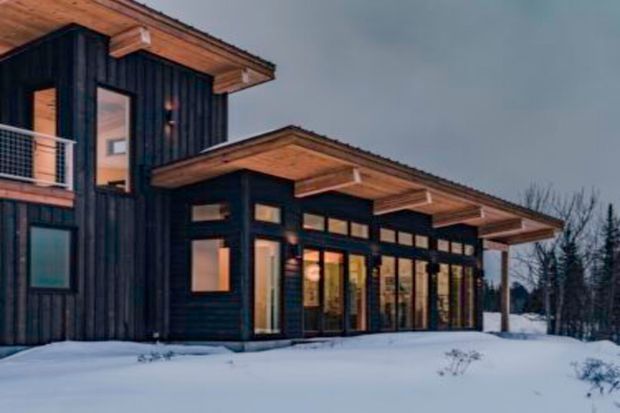 Exterior view of a contemporary multi-level building in a snowy setting, with dark vertical timber cladding, extensive glazing, projecting timber roof eaves, and a covered terrace or entry zone. Warm interior lighting contrasts with the cool dusk atmosphere and snow-covered landscape.
