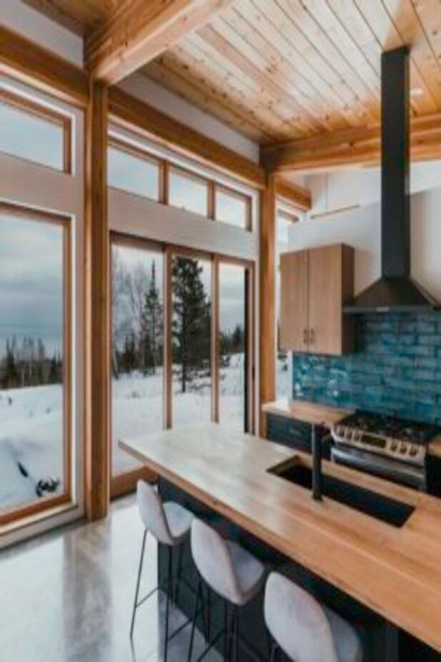 A contemporary kitchen in a timber-framed mountain setting, featuring a large light-wood island with three bar stools, black cooktop and sink, integrated cabinetry, a vivid blue tile splashback, and a black rangehood. Floor-to-ceiling windows overlook a snowy forest landscape. The ceiling has exposed timber beams and pale timber boards.