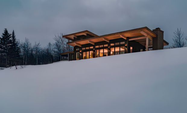 A modern single-storey house set within a snow-covered landscape at dusk. The building features extensive floor-to-ceiling glazing, strong horizontal roof overhangs, exposed timber-toned roof structure, dark framing, and a prominent masonry chimney. Warm interior lighting contrasts with the cool snowy setting and overcast sky.