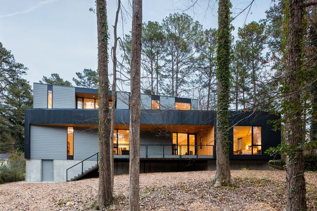 Exterior view of a contemporary multi-level house set among mature trees. The building has dark metal-clad projecting volumes, grey horizontal cladding, extensive warm-lit glazing, a raised deck or balcony, and a concrete lower level with an external stair.