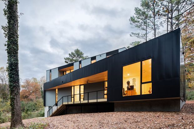 Exterior view of a contemporary multi-level house set among wooded landscaping. The building has a bold black metal-clad facade, angular roofline, extensive warm-lit glazing, projecting balconies, and dark metal railings. The house sits on a sloped, leaf-covered site beneath an overcast sky.