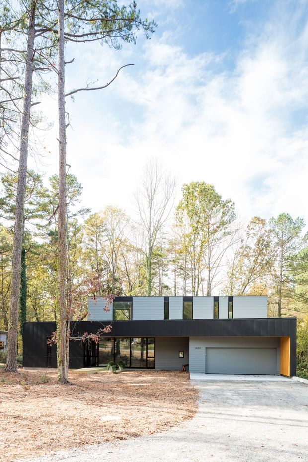 A modern two-storey house set among mature trees, with a long rectangular form, flat roof, dark horizontal lower-level cladding, light grey upper-level cladding, extensive glazing, and an integrated single garage. The front elevation is approached by a gravel driveway and surrounded by a wooded landscape.