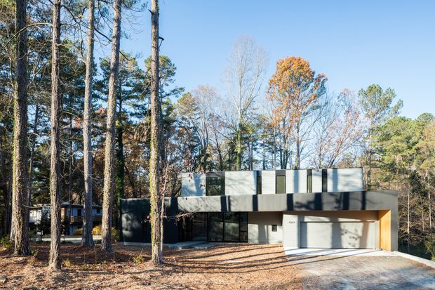 A modern two-storey residence set among tall mature trees, with a rectilinear form, flat roof, dark and light exterior panels, extensive glazing, and an integrated double garage. The home is surrounded by a wooded, sloping landscape under clear daylight.