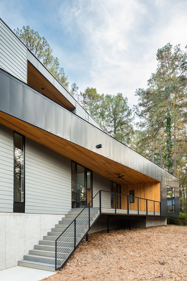 Contemporary two-storey residential facade set among mature pine trees. The building features horizontal light cladding, dark metal panel sections, warm timber soffits and recessed entry detailing, tall narrow windows, a raised timber-decked porch, and a concrete stair with black wire balustrade. The low-profile roofline and strong horizontal composition create a clean modern exterior.