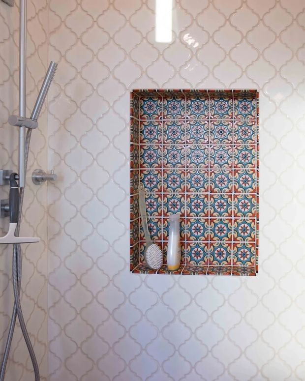 A close-up view of a shower area with white glossy interlocking quatrefoil-pattern wall tiles. A recessed niche features colourful patterned decorative tiles in blue, red, cream and grey, with toiletries inside. A chrome handheld shower rail and controls are visible on the left.