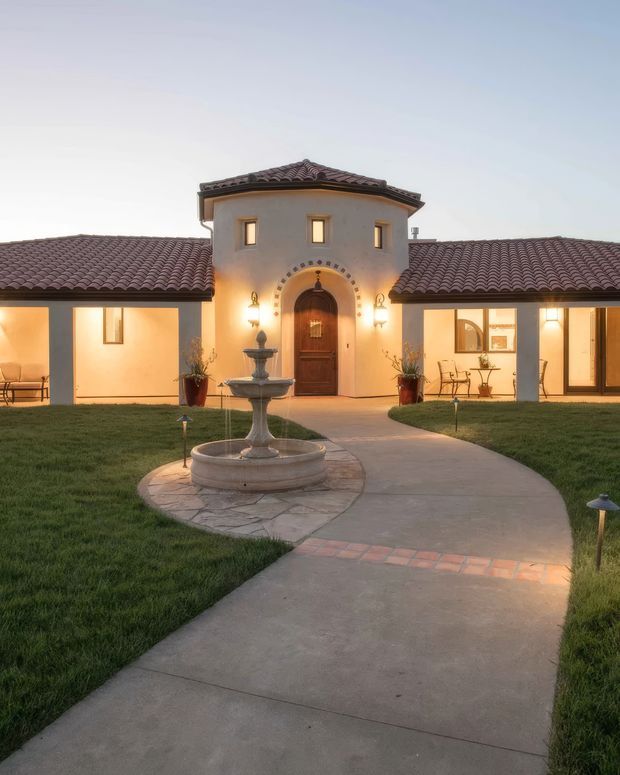 Front exterior view of a single-storey, symmetrical Mediterranean-style residence at dusk. The stucco facade has terracotta tiled roofs, a central two-storey entry tower with an arched timber front door and decorative stone surround, flanking wings with illuminated windows, and a curved concrete walkway leading through a lawn to a tiered stone fountain.