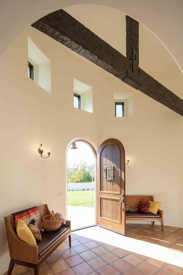 A sunlit, double-height entry hall with a rounded vaulted ceiling, exposed dark timber beams, small high-set windows, and a tall arched timber front door opening to a lawn. Terracotta square floor tiles, cream plaster walls, traditional wall sconces, and woven leather seating create a warm, rustic Mediterranean-inspired welcome space.
