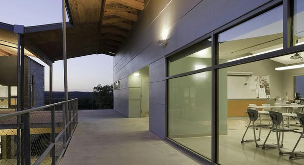 An elevated exterior walkway or covered terrace beside a modern institutional or commercial building. The view is dominated by a concrete-paved circulation space, dark metal-framed glazing, smooth grey wall panels, exposed timber roof structure, and metal balustrades. Through the large windows, a bright classroom or meeting room with tables, chairs, cabinets, and wall-mounted displays is visible. The building sits above a distant landscape and appears to have a contemporary, functional design.