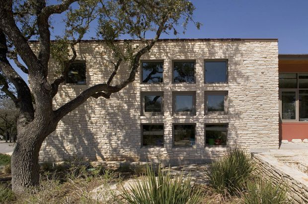 Exterior view of a contemporary low-rise building with a pale textured masonry facade and a regular grid of dark-framed windows. A mature tree and drought-tolerant landscape planting partially screen the facade, while a glazed connecting section is visible at the right.