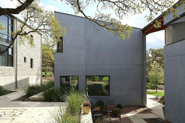 Exterior view of a contemporary multi-building residential complex arranged around a landscaped courtyard. The buildings feature smooth dark-grey rendered walls, pale brick masonry, large black-framed windows, and simple flat or shallow-pitched roof forms. Paved paths, gravel garden beds, ornamental grasses, mature trees, and a small outdoor seating area occupy the foreground.