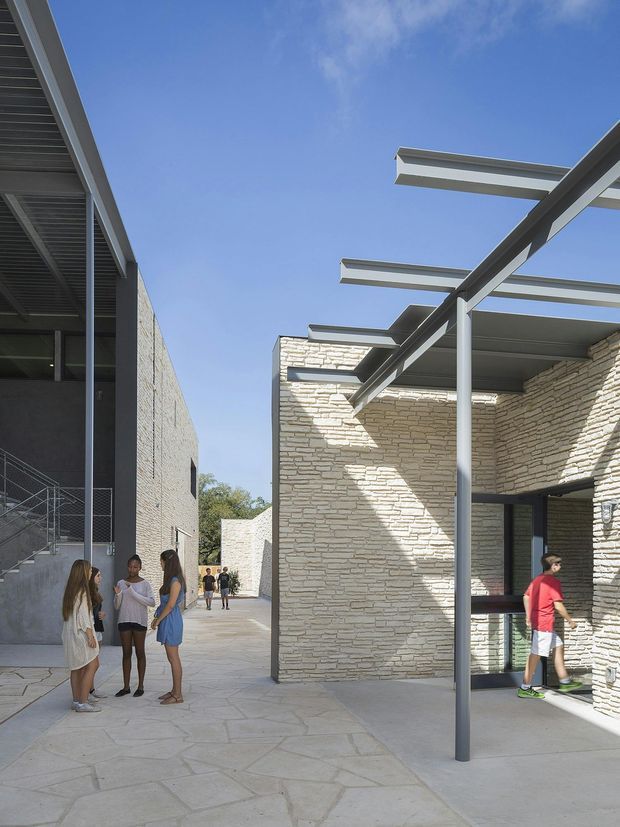 A sunlit contemporary institutional or commercial courtyard between low-rise buildings. Pale stacked-stone walls and broad concrete paving define the space, while dark metal-framed glazing and exposed steel pergola beams create strong linear shadows. People provide scale, and an external stair with metal railings is visible along the left building.