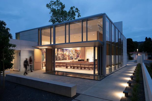 Contemporary two-storey glass-fronted building photographed at dusk, with extensive floor-to-ceiling glazing, dark metal framing, pale solid wall panels, and a projecting upper-level volume. Warm interior lighting reveals a spacious communal room with long bench seating and artwork, while a paved walkway and low concrete site wall frame the exterior.