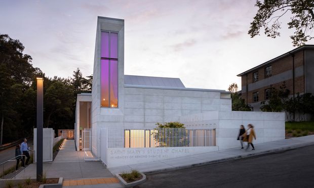 Exterior architectural photograph of a contemporary church or chapel at dusk. The compact white rendered or concrete building has a tall narrow illuminated vertical window tower with purple and warm amber lighting, a simple pitched metal roof, a perforated or vertical-bar fence, paved entry walkway, and restrained modern detailing. A larger brick building and trees surround the site.