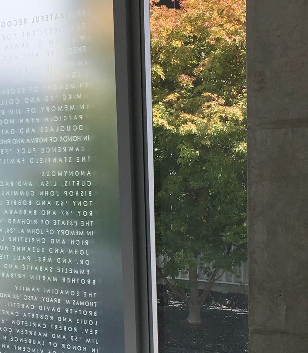 Close-up view of a tall glazed doorway or window with reversed white lettering printed on the glass, set within a dark, smooth concrete or stone wall. Through the glazing is a landscaped courtyard with a leafy tree and dark ground cover. The image appears to document an institutional or commercial architectural entrance, with the glass text and heavy wall framing as the main features.