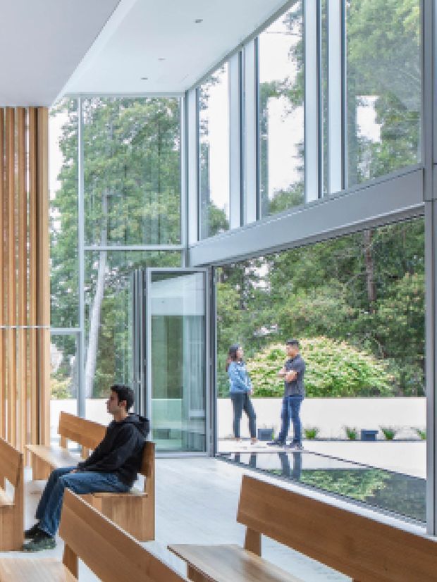 A bright contemporary double-height interior with extensive floor-to-ceiling glazing overlooking mature greenery. Warm vertical timber wall panelling and built-in timber bench seating contrast with the cool pale floor and exposed metal-framed glass doors and windows. An open glazed doorway connects to an outdoor terrace, while people occupy the space and terrace.