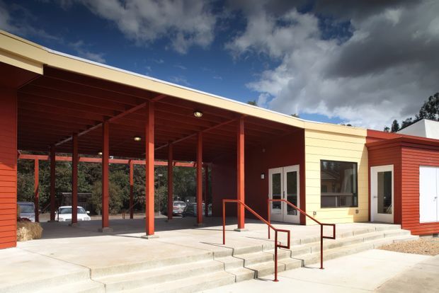 Single-storey contemporary building exterior with a broad red-painted covered entry or carport structure, pale yellow horizontal cladding, red vertical posts and trim, concrete steps and accessibility handrails, and a white garage-style door. The image is photographed from outside and also shows a landscaped, wooded setting beyond the covered area.