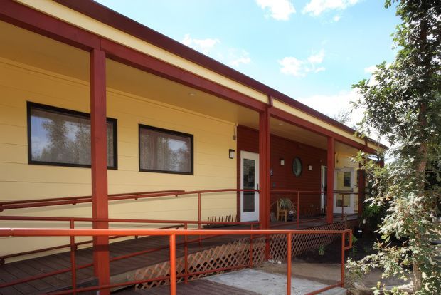Single-storey building exterior with a long covered veranda and accessible ramp leading to the entrance. The facade combines cream horizontal weatherboard-style cladding with dark red-brown posts, beams, railings and trim. Several windows and doors open onto the veranda, with mature planting along the right side under a bright blue sky.