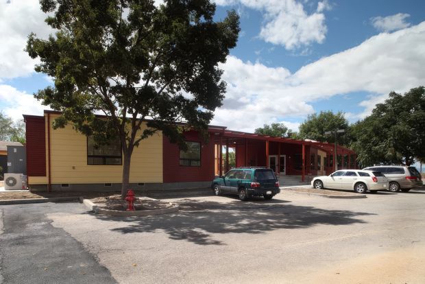 Single-storey contemporary low-rise building viewed from a paved parking area. The facade combines deep red vertical and horizontal elements with pale yellow-beige wall panels, dark-framed windows, and a covered entrance or walkway supported by red posts. Mature trees partially shade the building and several parked cars occupy the foreground.