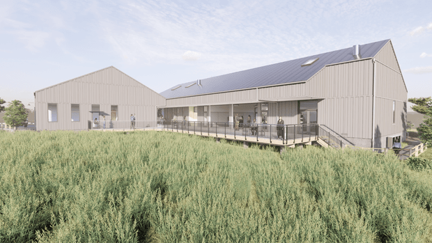 A rendered exterior view of a large contemporary agricultural-style building complex set behind tall grasses. The structures feature pale vertical metal cladding, simple gabled roof forms, dark grey standing-seam roofing, skylights, and extensive covered outdoor glazing and verandah decking. The image presents a low-rise, multi-wing building in a landscaped rural-looking setting.