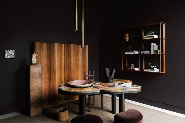A dark, refined dining area with a sculptural round dining table, upholstered stools, a tall walnut-toned storage cabinet, open timber shelving, and black walls. Two slim pendant lights hang above the table, while a textured neutral carpet and carefully arranged decorative objects add warmth and visual contrast.
