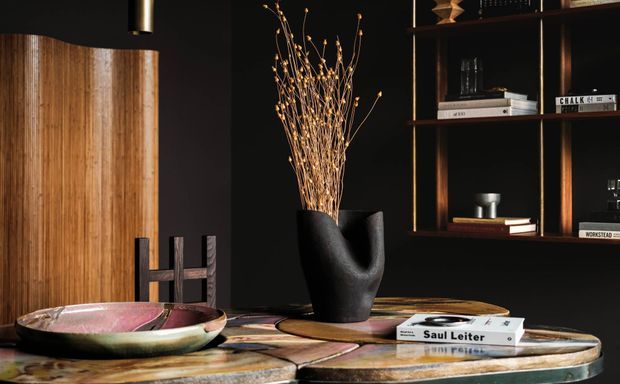 A moody, refined interior vignette featuring a dark timber and black built-in shelving unit, a sculptural black vase with dried branches, art books, and a shallow decorative bowl on a richly patterned stone or timber console. Warm wood tones and low lighting create a dramatic, sophisticated atmosphere.