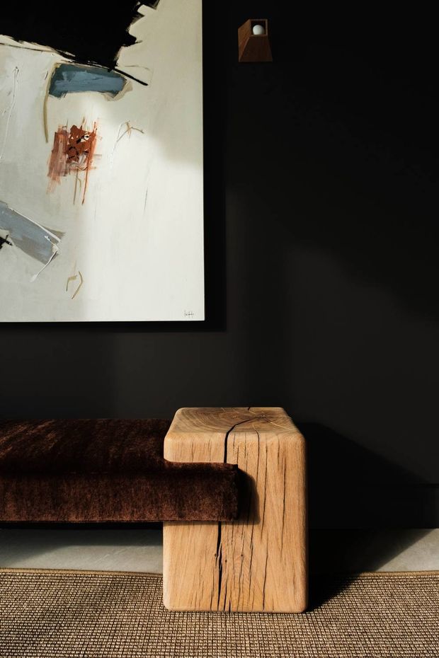 A close-up interior vignette featuring a dark brown upholstered sofa with a substantial sculptural timber end table or armrest integrated at its side. The room has matte black walls, a textured neutral woven floor covering, a small wall light, and a large abstract artwork with expressive black, cream, blue-grey, and rust marks. Strong directional sunlight creates dramatic shadows and highlights the timber grain.