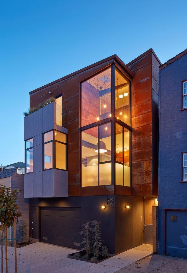 A narrow multi-storey urban townhouse facade photographed at dusk. The building combines dark rust-toned metal cladding with charcoal panels, extensive black-framed glazing, projecting box-like volumes, and a tall glazed vertical bay revealing warm interior lighting and pendant fixtures. A garage door and recessed entry sit at street level, with neighbouring attached buildings visible on either side.