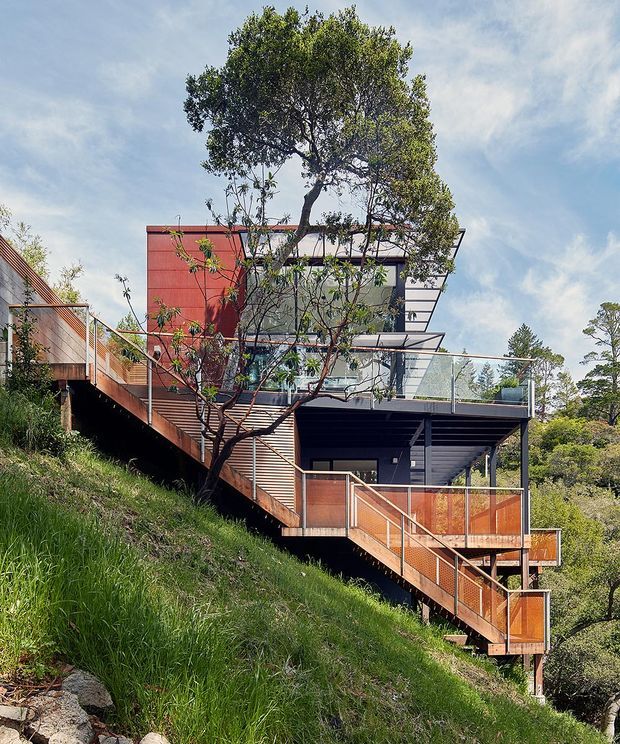A contemporary multi-level house stepping down a steep, grassy hillside. The building features dark horizontal cladding, a bold red panelled upper volume, extensive glazing, projecting flat roof planes, glass balustrades, and extensive timber decking and stairs with warm orange-toned screening. Mature trees and surrounding vegetation frame the elevated structure.