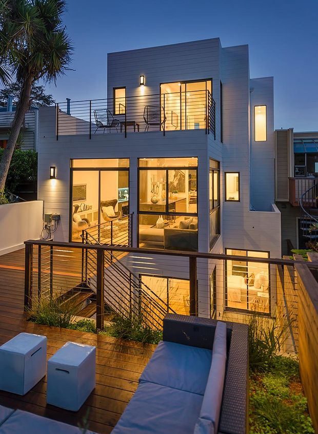 Twilight exterior view of a narrow, multi-level contemporary home with light horizontal cladding, extensive illuminated glazing, and layered timber decks. Black horizontal cable railings and stair balustrades define the upper terrace and connecting stairs. Outdoor seating and a lounge chair occupy the foreground deck, while warm interior lighting reveals living areas through the windows. A sculptural palm-like tree and neighbouring urban homes frame the scene.