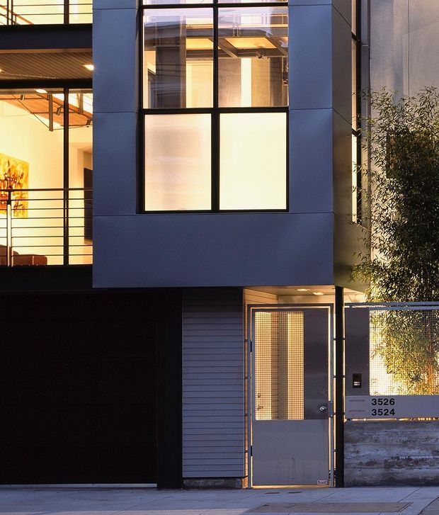 A contemporary multi-storey urban building facade viewed from the street, featuring dark blue-grey rendered wall panels, large black-framed glazed windows, a recessed entry door with wire-mesh glazing, a dark garage or service opening, and a metal pedestrian gate. Warm interior lighting is visible through the glazing, with a planted tree beside the entrance.