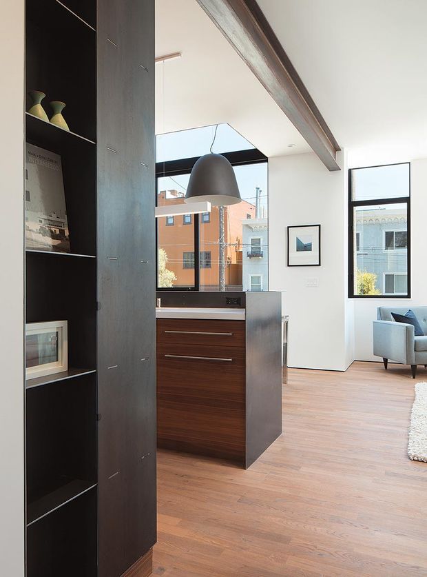 A bright contemporary open-plan interior featuring a compact kitchen island with dark wood drawer cabinetry and a dark stone benchtop, set against pale walls and warm timber flooring. A tall dark built-in shelving and storage partition occupies the foreground, while a large dark pendant light, exposed timber ceiling beam, black-framed windows, and a small seating area are visible beyond. Urban buildings can be seen through the glazing.