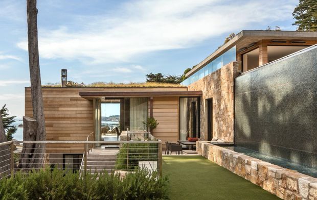 Contemporary multi-level residence set on a coastal, sloping site, with horizontal timber-clad walls, extensive glazing, flat rooflines, a planted roof section, and a large stone-clad water feature or pool wall. A terrace with metal balustrading and outdoor seating overlooks the ocean, visible through the glazing and beyond the property.