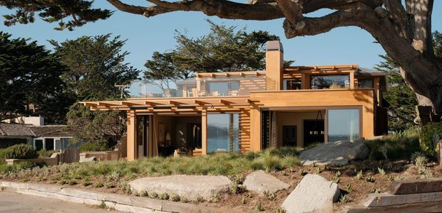 Exterior view of a contemporary two-storey residence set among mature coastal trees and landscaped planting. The house features warm timber-toned cladding and framing, expansive glazing, broad roof overhangs, a pergola-like timber structure, and a mostly flat roofline with a prominent chimney or vertical masonry element. Large natural boulders and grasses form part of the front landscape.