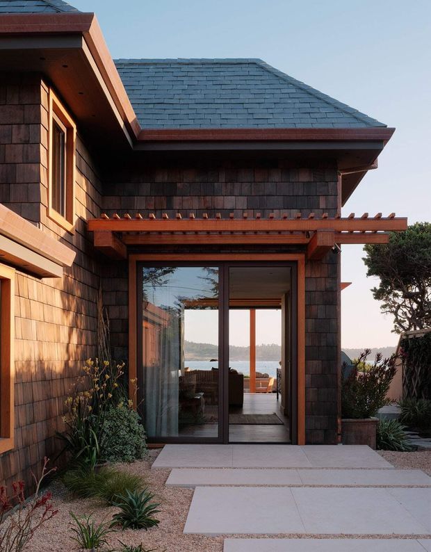 A close exterior view of a dark cedar-shingled coastal-style house, showing a projecting roof with slate-like tiles, timber-framed glazing, and a timber pergola above a glazed entry or living-area doorway. Concrete stepping slabs lead through a landscaped gravel garden with drought-tolerant planting. Water and a wooded shoreline are visible beyond the interior glazing.