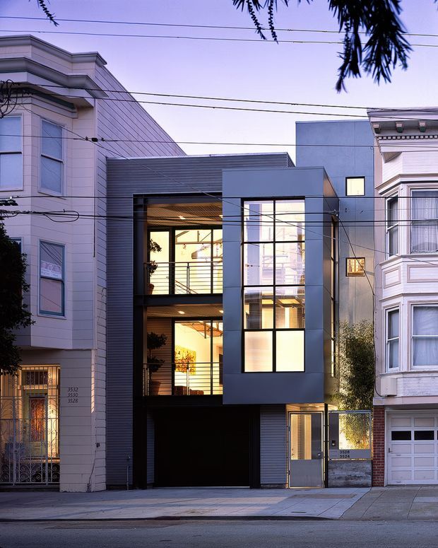 Street-facing view of a narrow contemporary multi-storey urban residence inserted between traditional neighbouring homes. The facade combines dark horizontal cladding, smooth grey panels, extensive glazed openings, and a recessed ground-level garage or entry. Traditional bay-fronted houses flank the sharply modern central building, with overhead utility wires crossing the view.