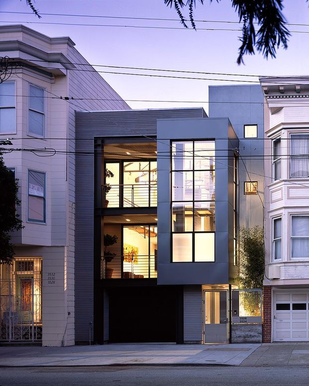 Street-facing photograph of a narrow contemporary multi-storey urban residence inserted between traditional neighbouring buildings. The facade combines dark horizontal cladding, smooth grey panels, large glazed openings, recessed balconies, and a ground-level garage and entry. Utility wires cross the view, with warm interior lighting visible through the windows at dusk.