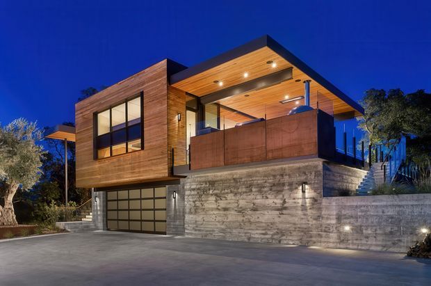 Night-time exterior view of a contemporary multi-level residence on a sloped site. The house combines warm timber-clad upper volumes, broad dark-framed glazing, exposed concrete retaining walls, a recessed garage, and a large covered upper terrace with timber soffit and integrated lighting. Landscaped planting and an exterior stair are visible around the building.
