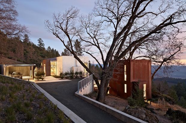 A modern hillside residence viewed from an elevated exterior approach at dusk. The composition shows flat-roofed, rectilinear buildings integrated into a sloping, wooded site, with pale rendered walls, extensive glazing, and a prominent rust-red metal-clad volume. A large leafless tree frames the foreground, while a paved driveway and pedestrian ramp with metal balustrades lead toward the house. Distant mountain or valley views are visible beyond the property.