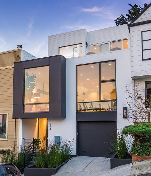 Street-facing view of a narrow multi-storey contemporary townhouse with a strong geometric facade. The home combines white horizontal cladding, dark charcoal projecting window bays and garage door, extensive glazing, and a recessed illuminated entry. Tall built-in planter beds and a sloping concrete driveway frame the frontage.