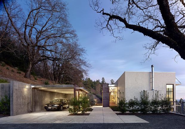Exterior view of a modern single-storey residence set into a wooded, sloping site. The composition shows a minimalist concrete or rendered building with flat rooflines, large vertical glazing, a covered integrated carport, paved entry path, landscaped planting, and a visible railing at the edge of the property. The image is taken at dusk with warm interior and exterior lighting against a cool blue sky.