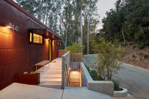 Exterior view of a compact contemporary dwelling set among dense trees, with a dark rust-coloured metal-clad facade, warm recessed lighting, projecting windows, and the address number 441. A paved entry path and raised landing lead to the entrance, alongside a concrete sunken stairwell or lightwell with integrated planting and metal balustrade. A narrow driveway curves along the wooded site.