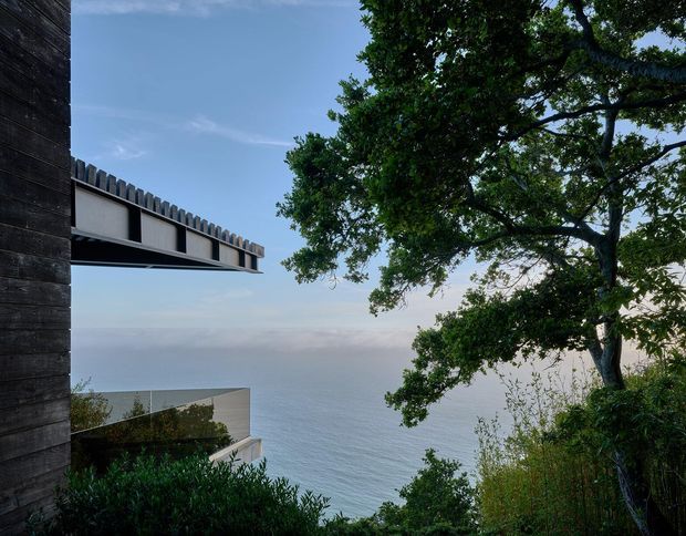 Exterior architectural view of a modern coastal residence set above the ocean. The image shows dark vertical timber cladding at left, a projecting roof or balcony edge with a serrated parapet detail, and a lower pale concrete or rendered volume partially visible through dense coastal vegetation. The ocean and horizon form the background, framed by mature trees and landscaped planting.